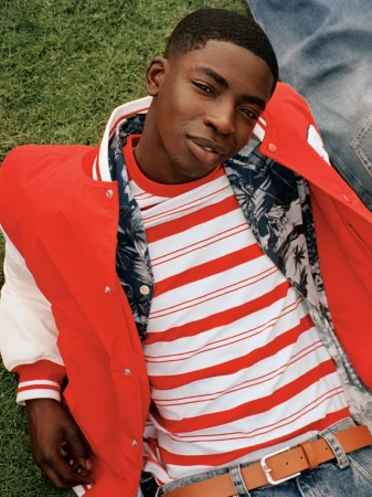 Young Black man in an open varsity jacket and striped shirt poses on a green lawn with denim-clad legs in the background.