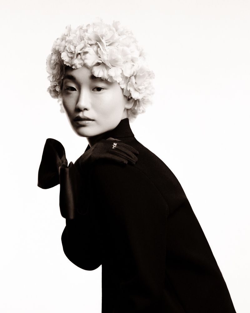 Headshot of a young Asian woman wearing a flower headdress, black turtleneck, and opera gloves against a bright white backdrop.