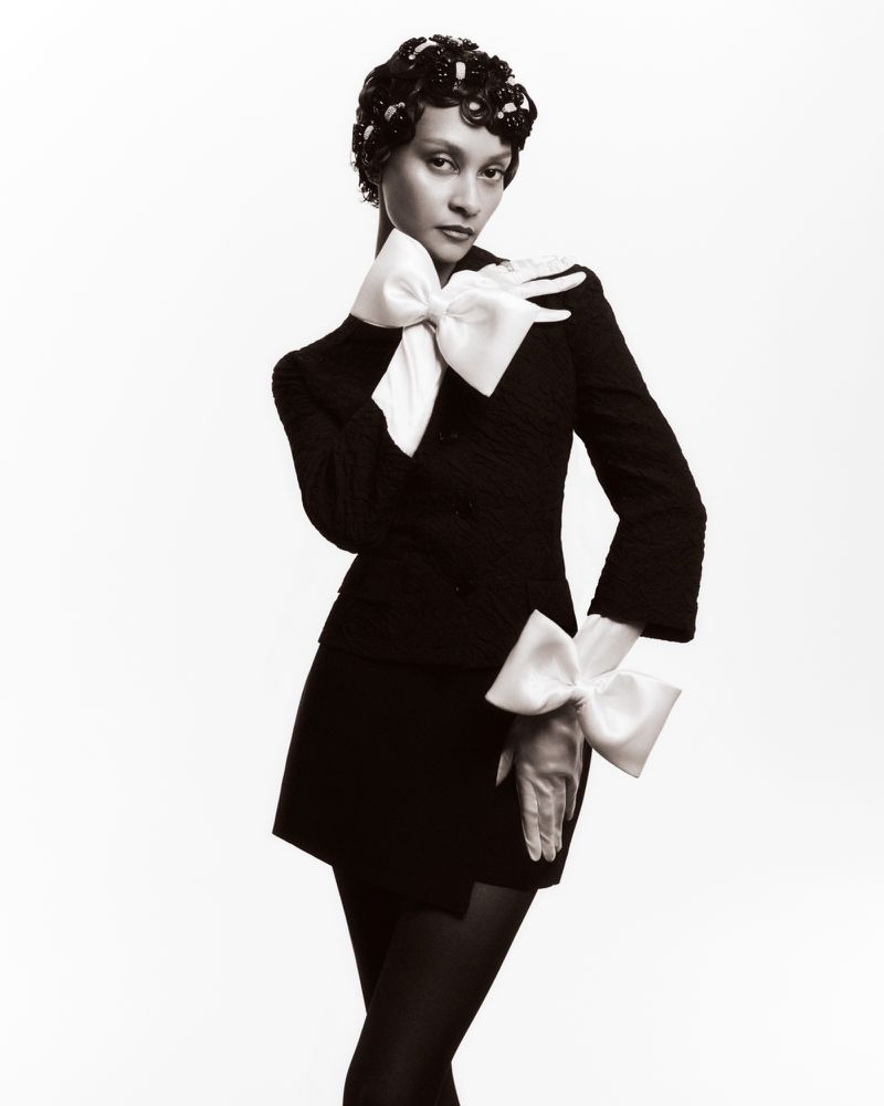 Portrait of an adult woman wearing a black dress with oversized white bows on the sleeves, posing against a white backdrop in a studio.