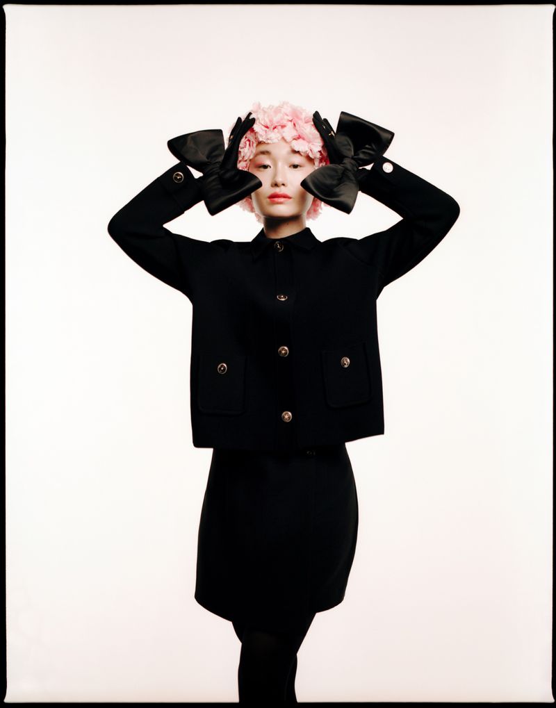 A stylish Asian woman with a pink flower headpiece, wearing a black jacket, skirt, gloves with bows, and posing in a white studio.