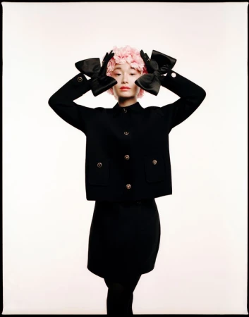 A stylish Asian woman with a pink flower headpiece, wearing a black jacket, skirt, gloves with bows, and posing in a white studio.