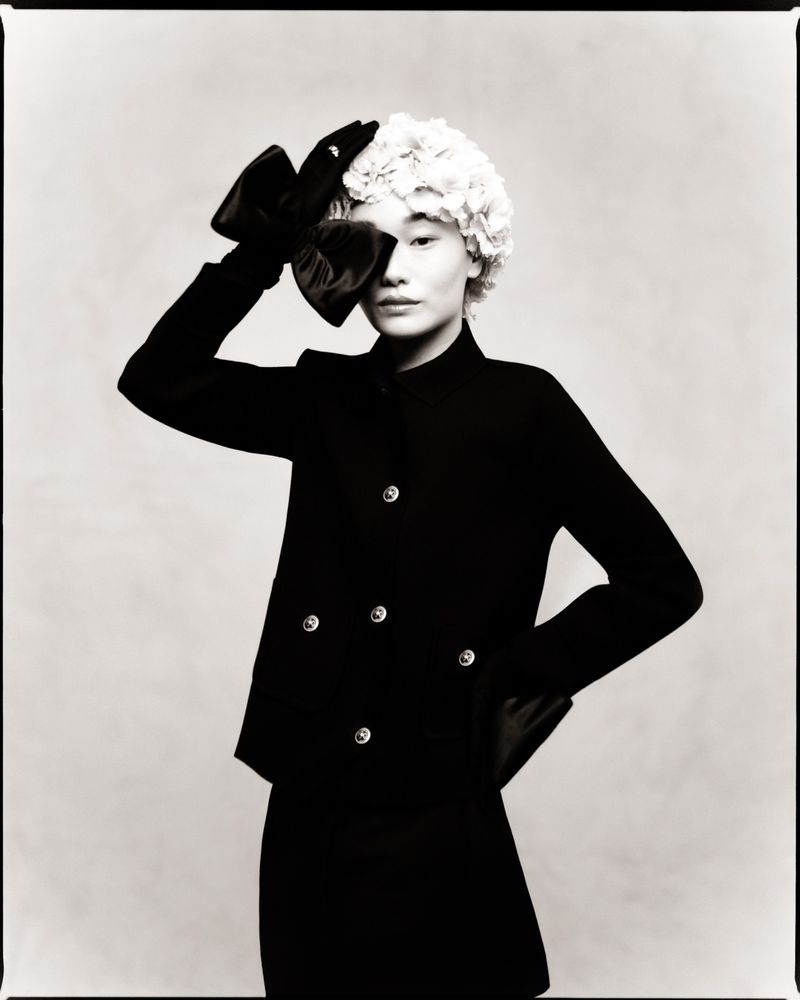 Black and white studio portrait of an Asian female fashion model wearing a black skirt suit and holding a large bow on her head.