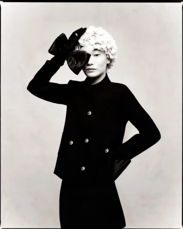 Black and white studio portrait of an Asian female fashion model wearing a black skirt suit and holding a large bow on her head.