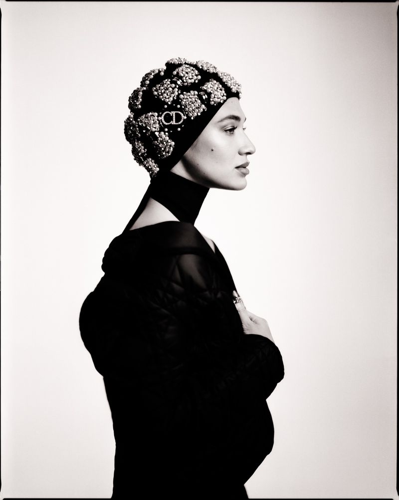 Black and white portrait of a stylish woman in a black outfit and a beaded head covering with a CD logo, against a plain white backdrop.