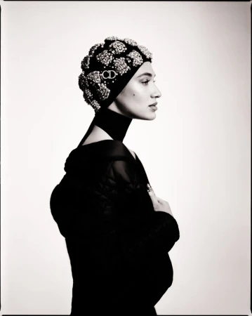 Black and white portrait of a stylish woman in a black outfit and a beaded head covering with a CD logo, against a plain white backdrop.