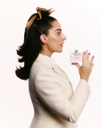 Elegant adult female in a cream suit presents a Miss Dior perfume bottle against a bright white backdrop. Fashion, beauty, and luxury.