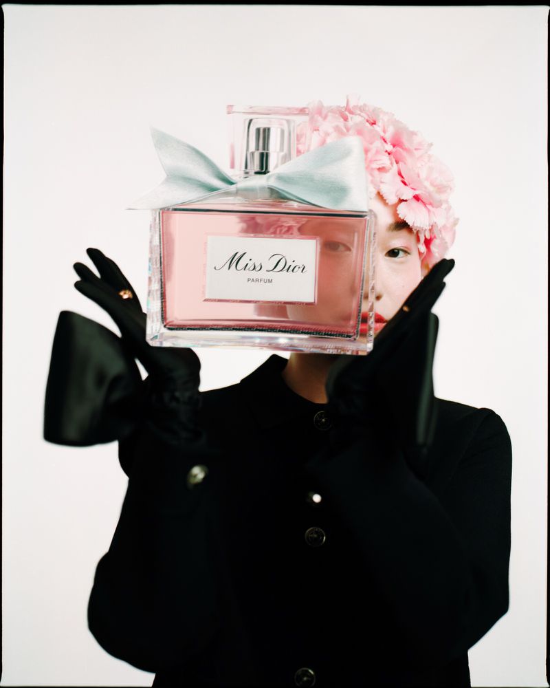 A person holds a large pink Miss Dior perfume bottle in front of their face, adorned with flowers, wearing a black jacket and gloves.