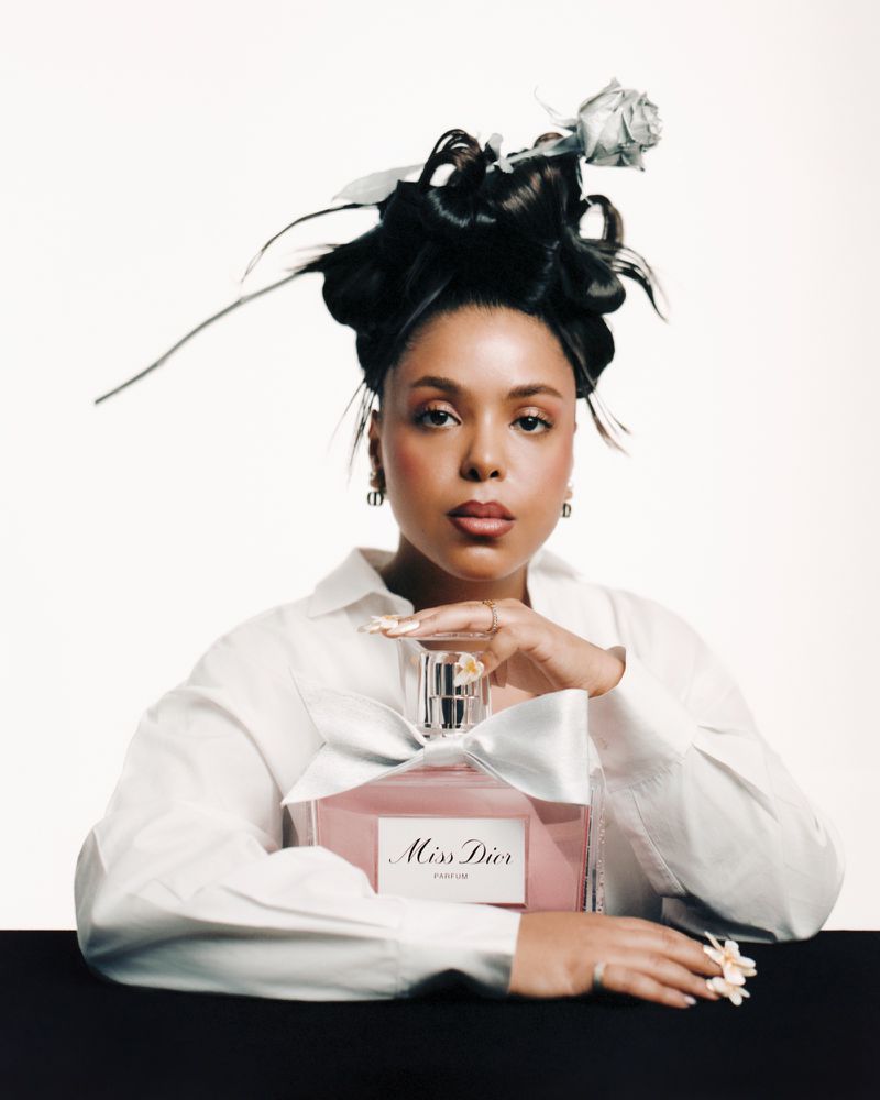 Glamorous beauty shot of an adult woman in a white shirt posing with a bottle of Miss Dior perfume against a white and black backdrop.