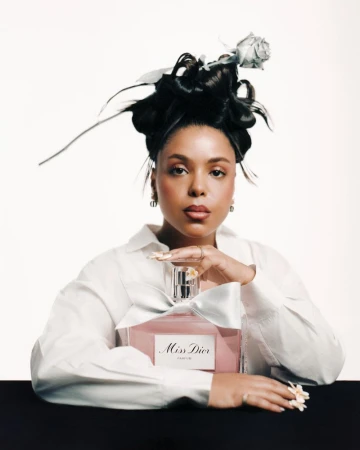 Glamorous beauty shot of an adult woman in a white shirt posing with a bottle of Miss Dior perfume against a white and black backdrop.
