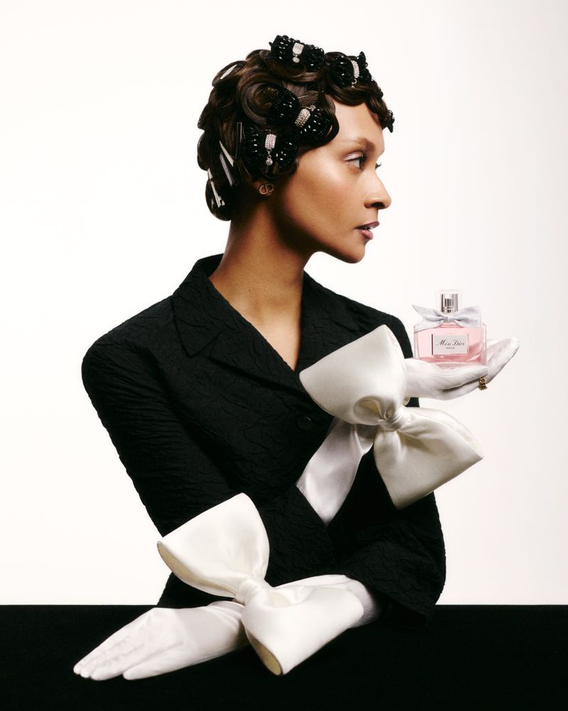 Elegant woman in a black textured jacket, white gloves with bows, and stylish hair, holding a Miss Dior perfume bottle against a white backdrop.