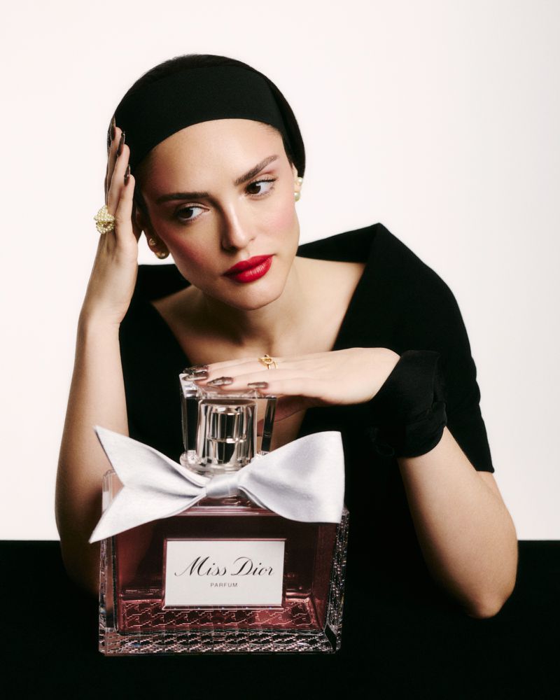Beauty shot of a woman with dark hair and red lipstick, posing with a bottle of Miss Dior perfume against a neutral backdrop.