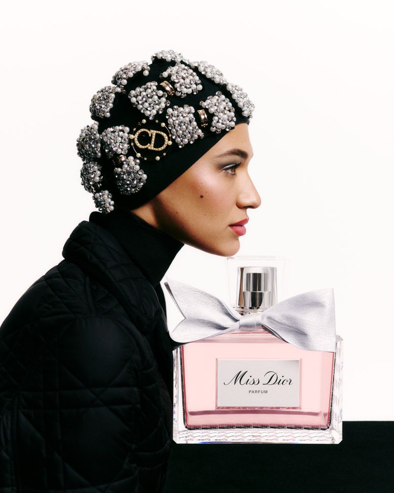 Close-up of a woman in an embellished beanie with a perfume bottle. The bottle has a silver bow against a bright studio backdrop.