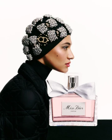 Close-up of a woman in an embellished beanie with a perfume bottle. The bottle has a silver bow against a bright studio backdrop.