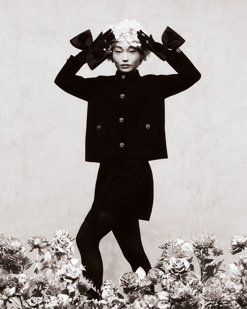 Monochrome fashion portrait of an Asian model in a black jacket, mini skirt, black tights, gloves with bows and a floral headpiece.