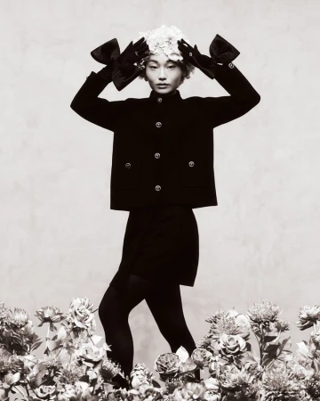 Monochrome fashion portrait of an Asian model in a black jacket, mini skirt, black tights, gloves with bows and a floral headpiece.