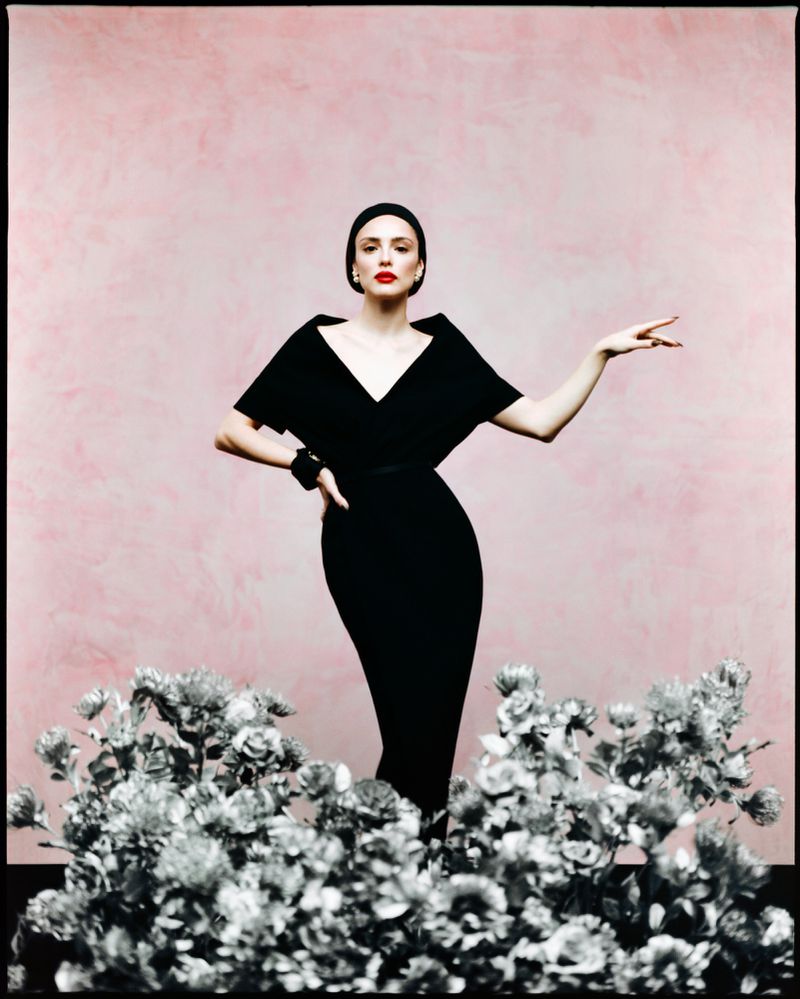 Elegant woman in black dress and headscarf poses in front of pink wall with flowers, fashion editorial photography. 