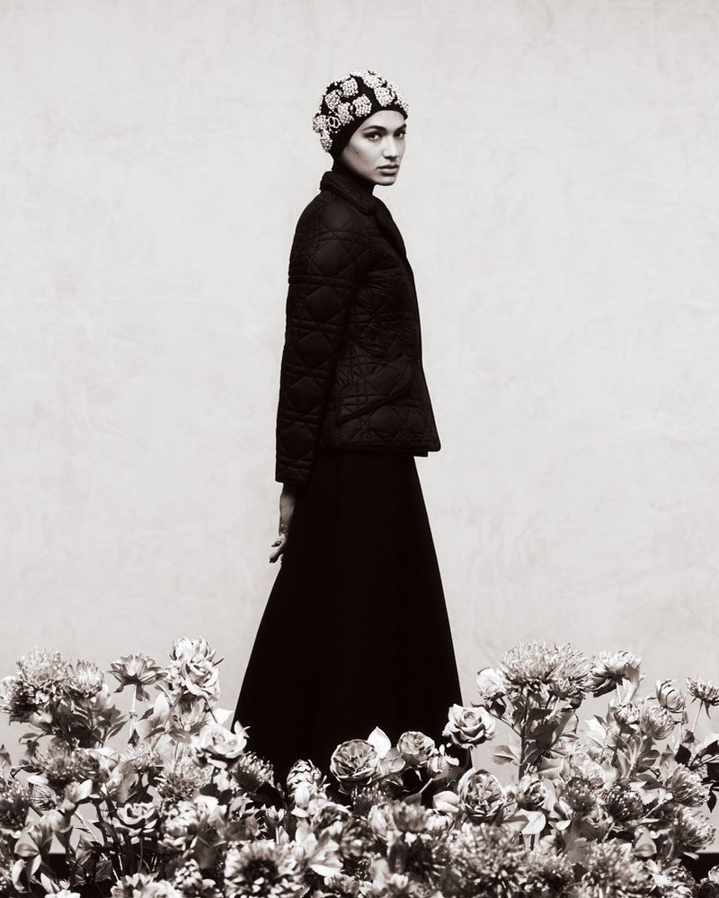 Elegant portrait of a woman in a black quilted jacket, long skirt, and beaded headpiece, standing among flowers against a neutral backdrop.