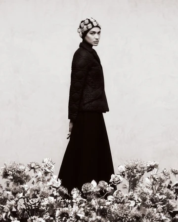 Elegant portrait of a woman in a black quilted jacket, long skirt, and beaded headpiece, standing among flowers against a neutral backdrop.