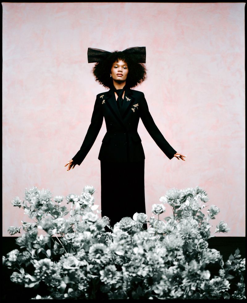 Fashionable woman in black suit with large bow, standing among white flowers against a pink background in a studio setting.
