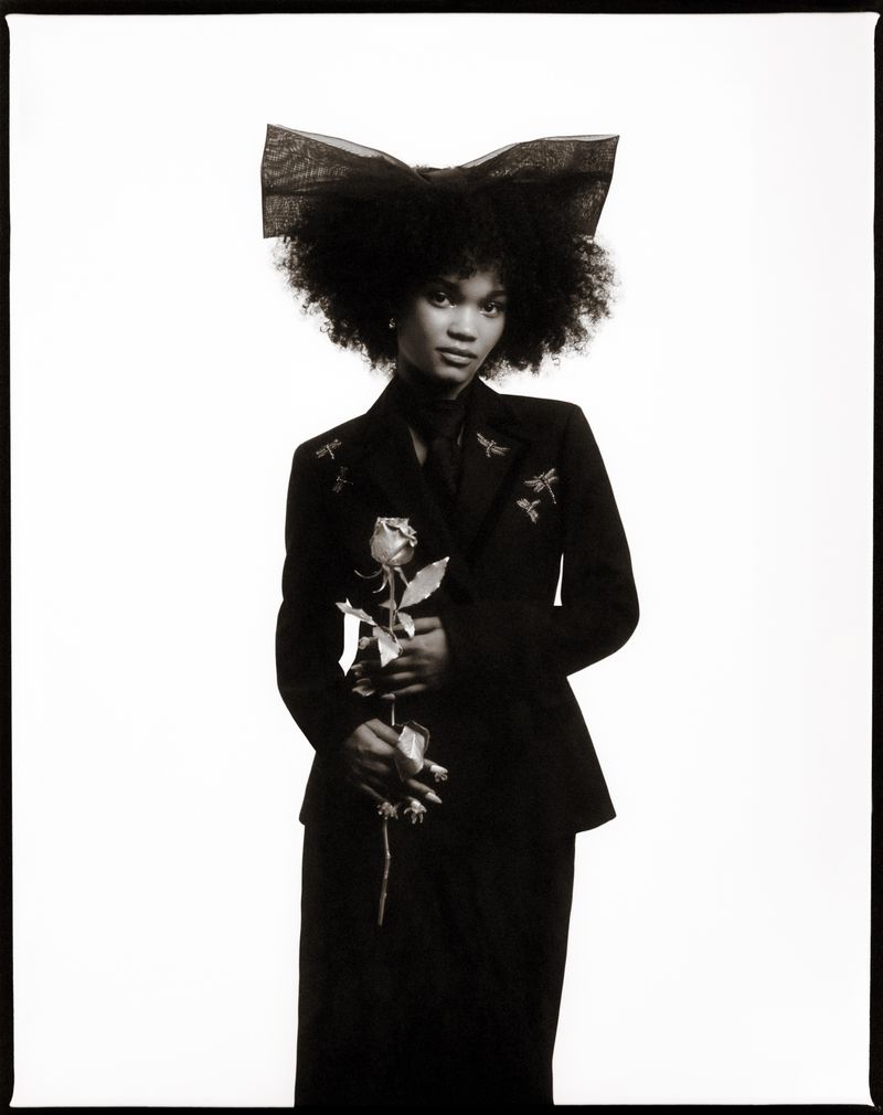 Portrait of a woman wearing a black jacket and a large bow in her hair, holding a rose against a white background