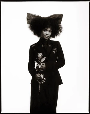 Portrait of a woman wearing a black jacket and a large bow in her hair, holding a rose against a white background