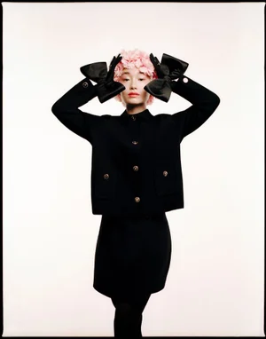 Model in black jacket with black bows and pink floral headpiece against a plain white background for Dior Beauty