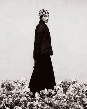 Model in a dark quilted jacket and embellished cap posing behind a dense arrangement of flowers in a monochrome photograph