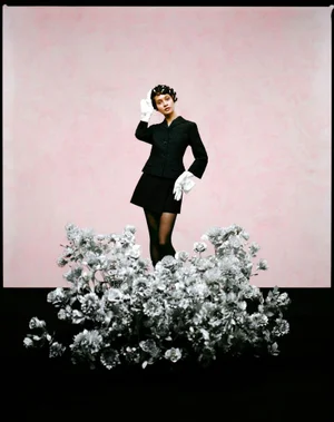 Woman in a black jacket and white gloves posing behind a large cluster of metallic silver flowers against a pale pink backdrop.