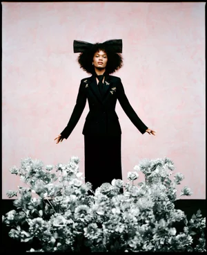 Model wearing a black suit and large bow headband standing behind silver flowers for Miss Dior campaign