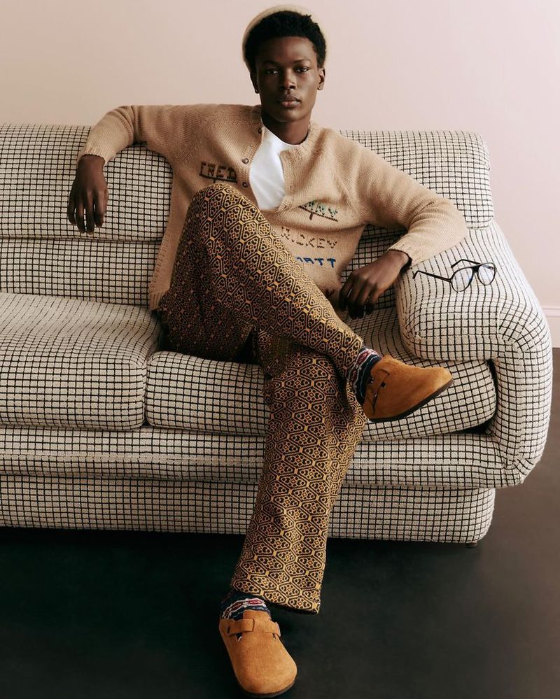Dark-skinned adult male model sits on a checkered couch wearing a beige cardigan and patterned pants in a studio with a pink wall.