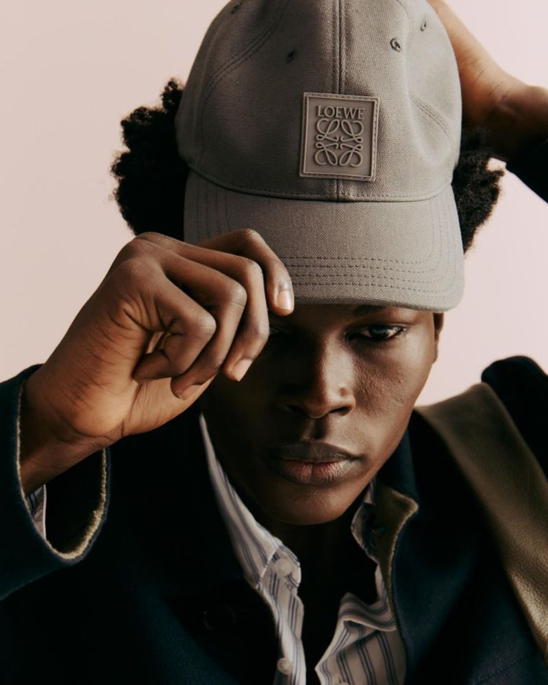 Portrait of a young Black man wearing a Loewe baseball cap and blue blazer against a light peach background, fashion photography.