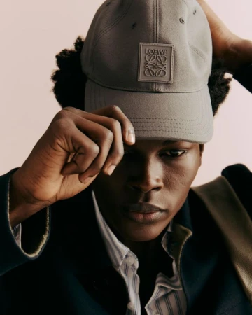 Portrait of a young Black man wearing a Loewe baseball cap and blue blazer against a light peach background, fashion photography.