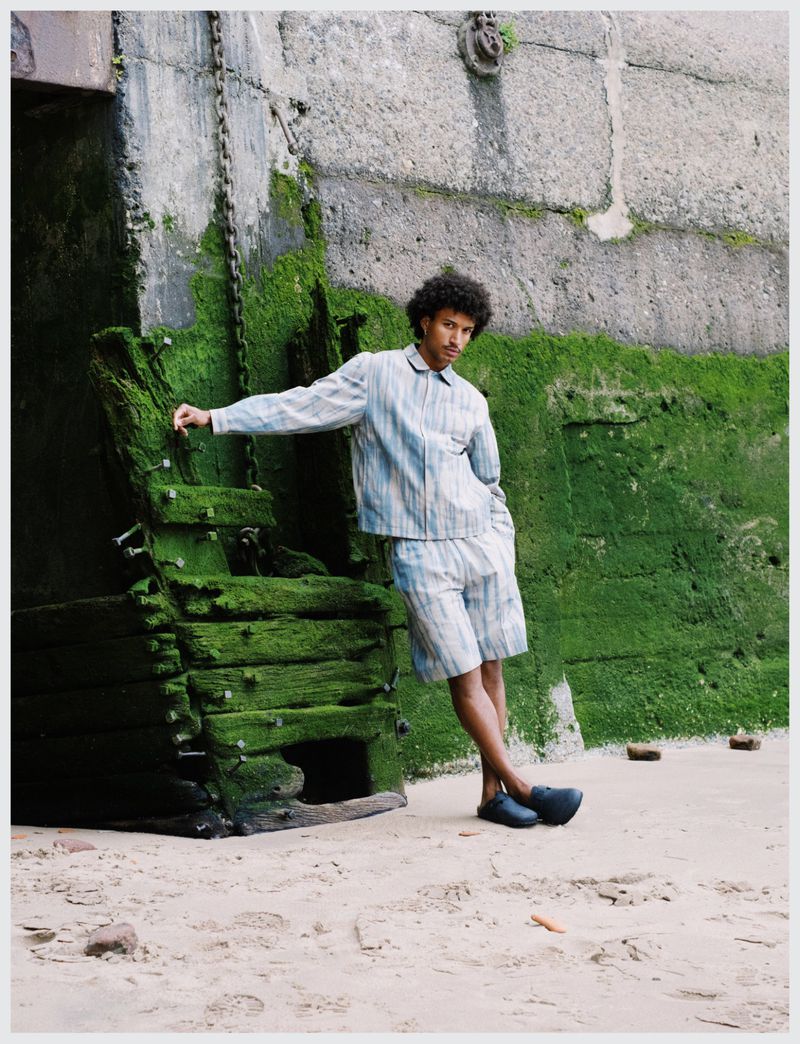 A young adult male in a striped shirt and shorts poses near a mossy stone structure on a sandy surface in an outdoor setting.