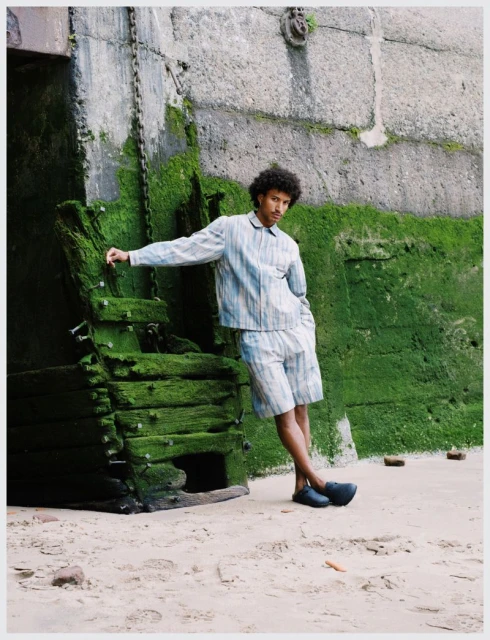 A young adult male in a striped shirt and shorts poses near a mossy stone structure on a sandy surface in an outdoor setting.