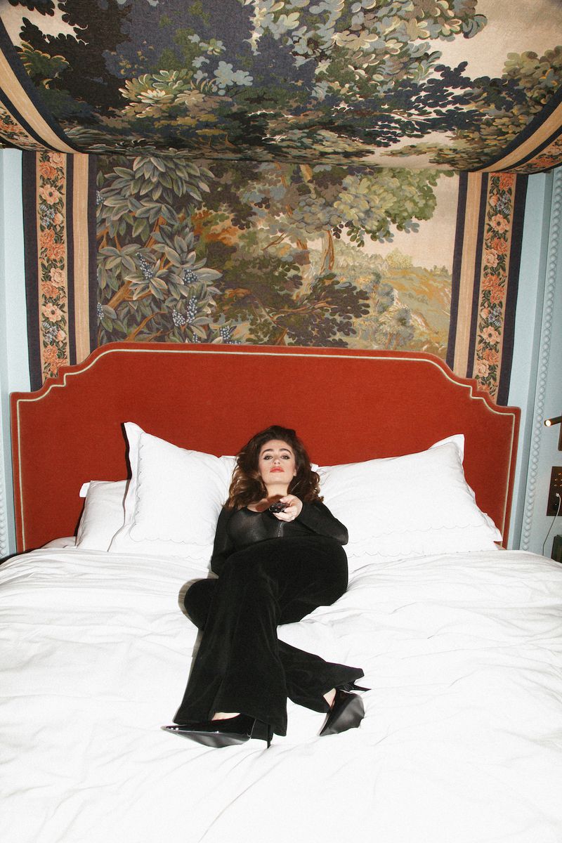 A woman relaxes on a bed in black clothing while using a remote control, surrounded by a white comforter, red headboard, and mural wallpaper.