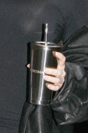 Hand holds a stainless steel travel cup with a metal straw. The cup has a Balenciaga logo on it. Person wears black clothing.