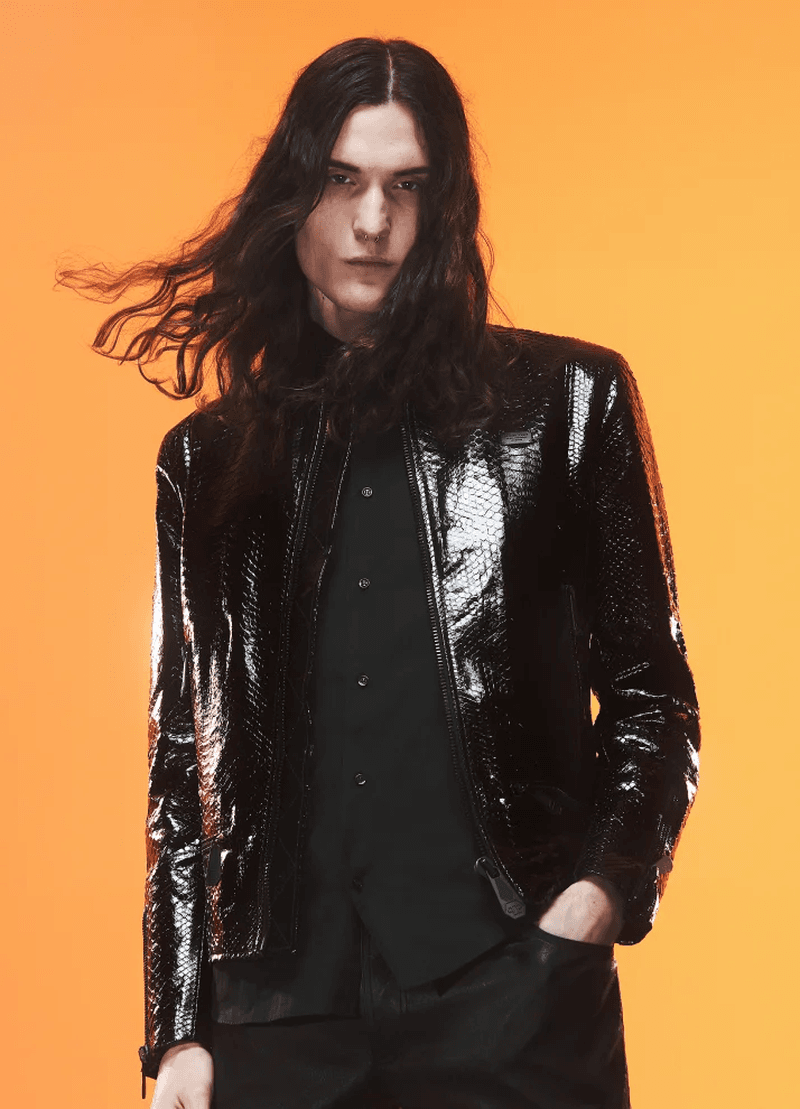 A portrait of a stylish adult male with long dark hair, wearing a shiny reptile-print jacket against a vivid orange background.