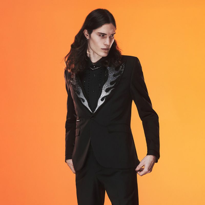 Man in a black suit jacket with a flame design posing against a graduated orange background in a studio setting for a fashion shot.