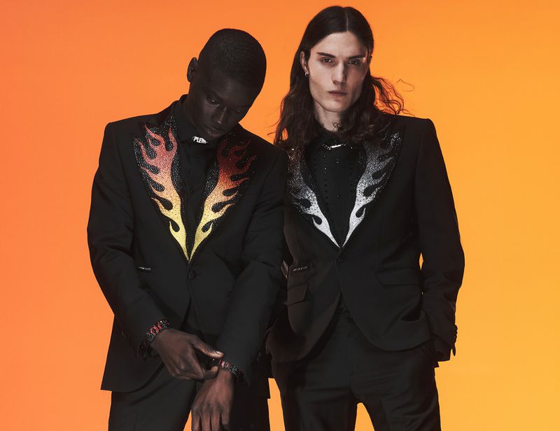Two male models pose in black suits with flame detailing on an orange gradient background. Fashion editorial shot.