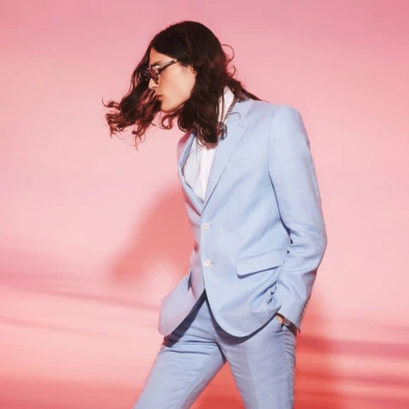 Man in a light blue suit and glasses poses against a pink backdrop with a wavy shadow pattern, creating a retro, fashion-forward look.
