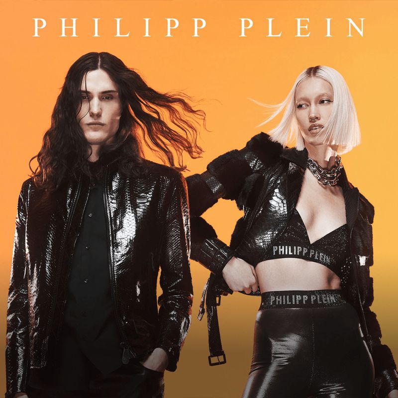 Two fashion models, an adult male and an adult female, are posing in black leather jackets with an orange gradient background in a studio.