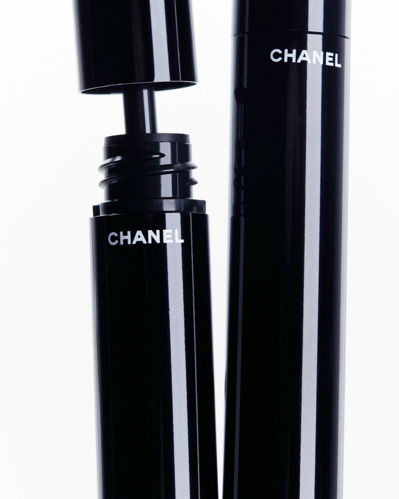 Black Chanel mascara tubes with white branding on a clean white background. One tube open with applicator visible. Beauty, makeup, product shot.