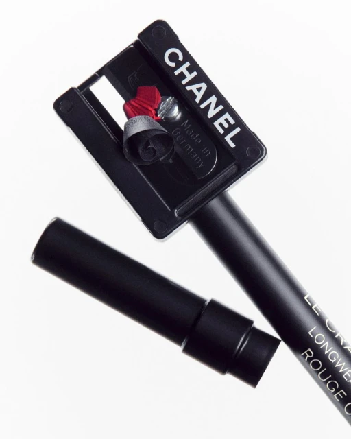 A close-up studio shot shows a black Chanel eyeliner pencil with its sharpener revealing a red eyeliner shaving on a white backdrop.
