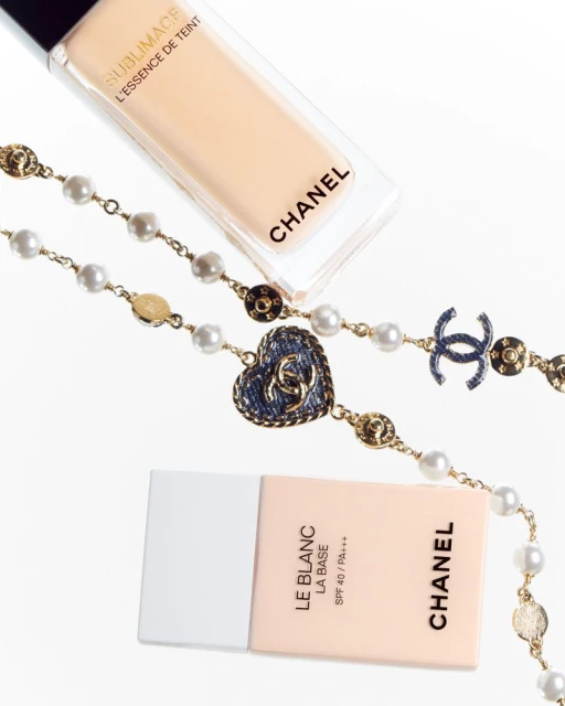 Luxury beauty product flat lay features Chanel foundation bottles alongside a gold and pearl Chanel necklace on a white background.