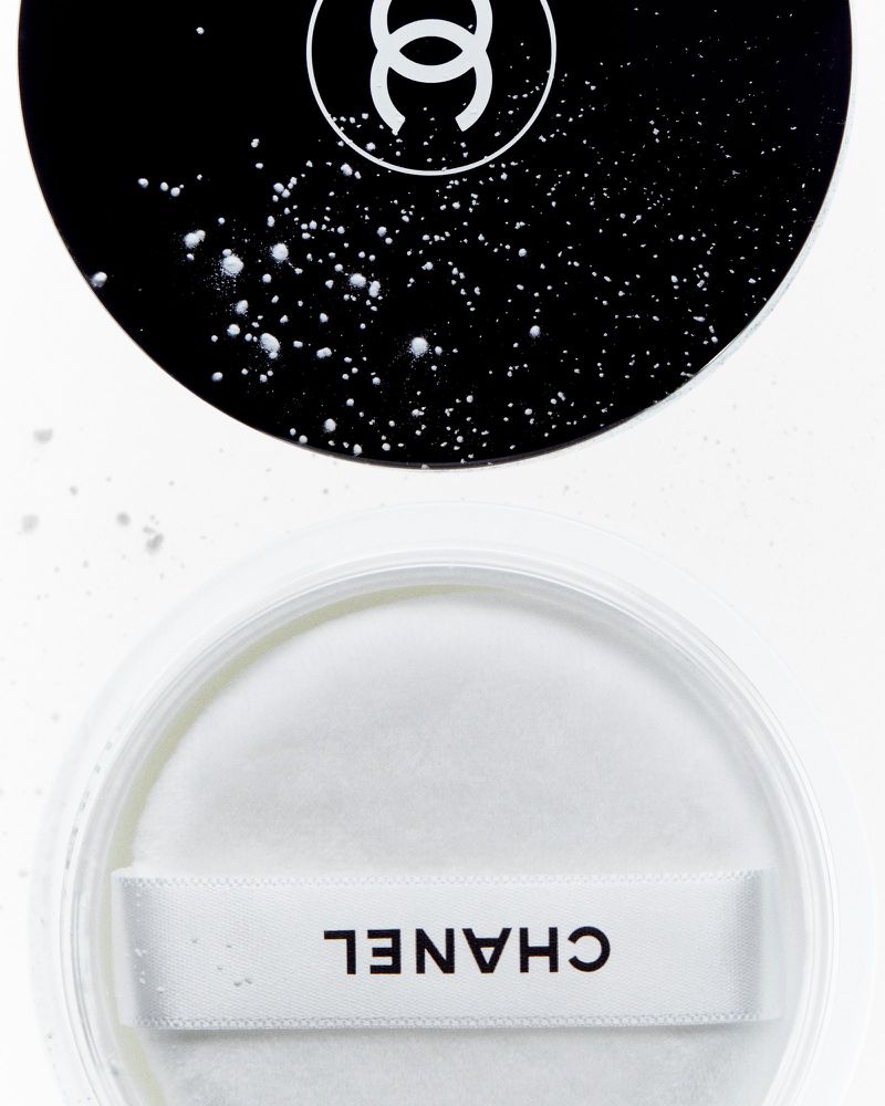 High-angle close-up of a CHANEL compact with white powder and a puff, featuring a black and white logo on a reflective surface.