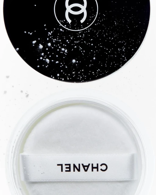 High-angle close-up of a CHANEL compact with white powder and a puff, featuring a black and white logo on a reflective surface.