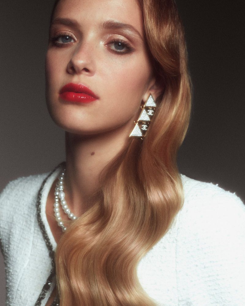 Close-up of a stylish adult woman with wavy blonde hair, pearl necklace, and triangle earrings, wearing a white tweed jacket.