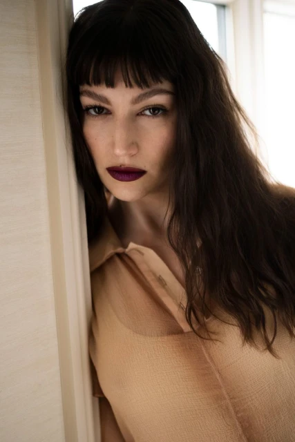 Close-up portrait of a woman with dark hair and bangs, wearing a tan collared shirt, leaning against a white wall near a window.