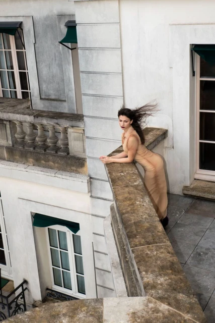 A woman in a long gown leans on a stone balcony overlooking a white building with windows and awnings in an outdoor urban setting.