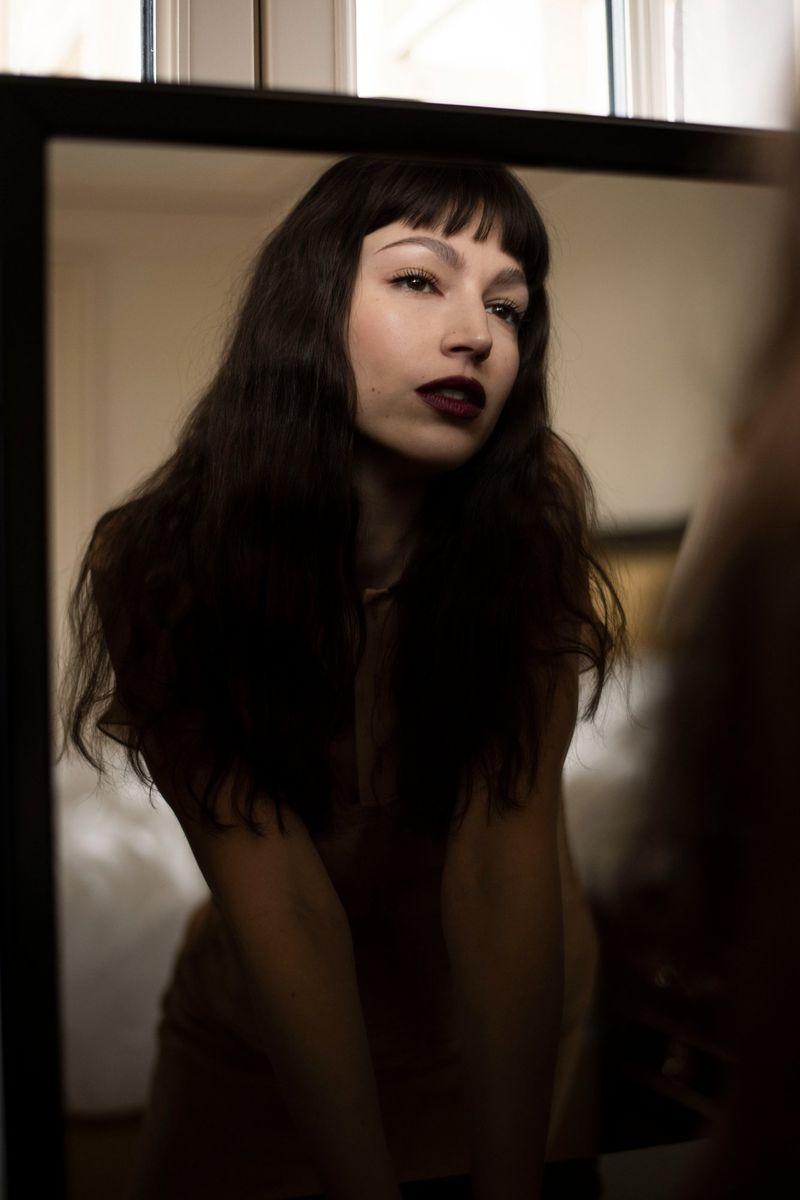 Portrait of a brunette woman with dark lipstick reflected in a mirror indoors, with a neutral color palette and soft lighting.